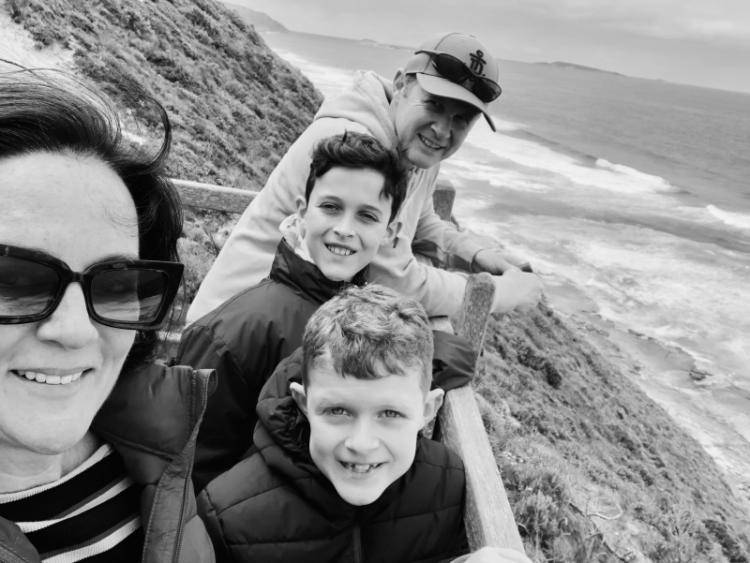 Kylie and her windswept family (two tween boys and husband, Stephen), smile at the camera while standing at a wintery beach lookout. Photo in greyscale. 